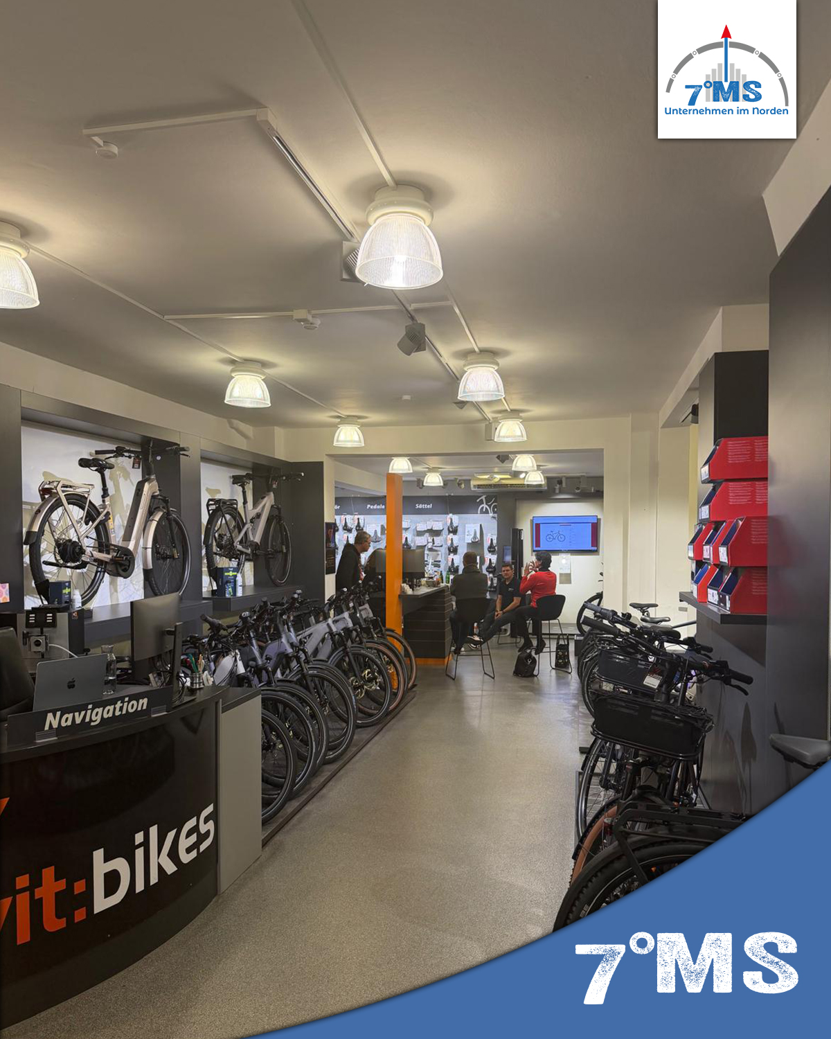 Im Fahrradgeschäft sind mehrere E-Bikes und Fahrräder auf Ständern ausgestellt. An der Theke steht ein Mitarbeiter, während im Hintergrund Kunden an einem Tisch sitzen und sich unterhalten. Der Bereich ist hell erleuchtet und modern gestaltet.