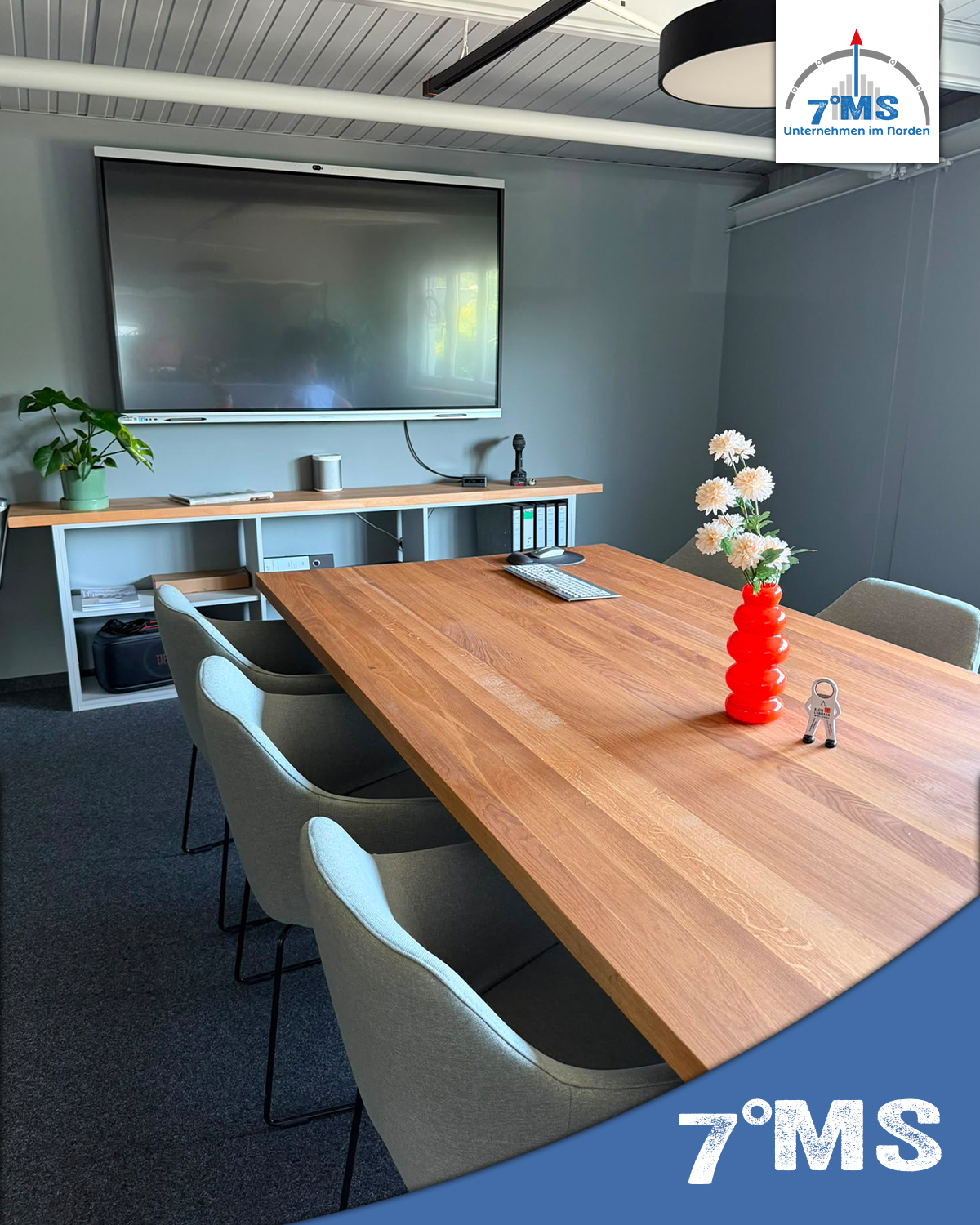 Ein moderner Besprechungsraum mit einem großen Holztisch, umgeben von grauen Stühlen. An einem Ende steht eine rote Vase mit Blumen. An der Wand hängt ein Bildschirm, neben dem ein Mikrofon und ein Laptop stehen. Das Raumdesign ist sachlich und einladend.